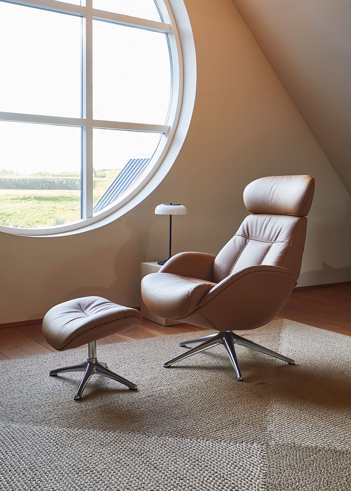 Elegant Lænestol med fodskammel i læderet Dakota Modern Cognac Brown med ben i poleret aluminium foran et rundt vindue